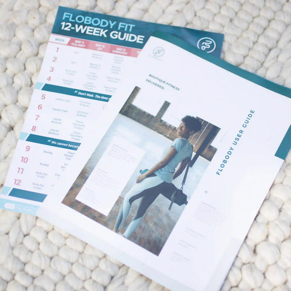 Two Flobody Fit 12-Week Guide fitness planner pages laid flat on a white textured knit blanket. The top page shows a teal header and workout schedule columns, while the bottom page is mostly white with a photo of a woman and the title 'FLOBODY USER GUIDE'.